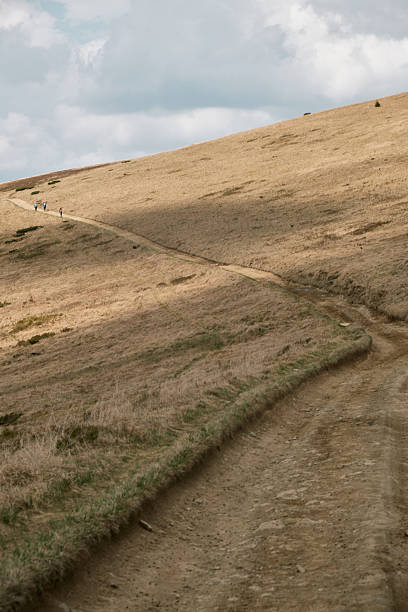 Solitary hiker ascending a trail through the rugged terrain of Svydovets mountain range in western Ukraine, under a partly cloudy sky.