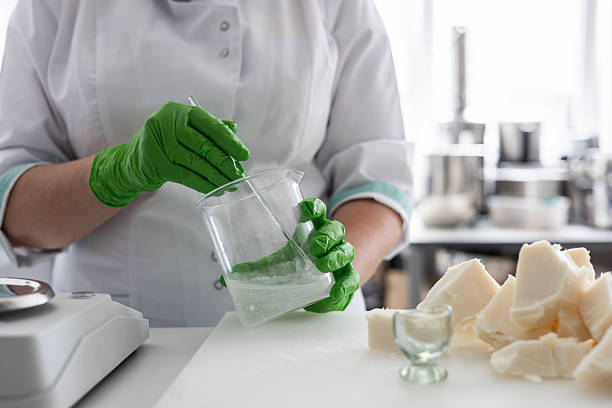A laboratory technician in a white coat and green nitrile gloves stirs a liquid in a beaker. raft production of organic cosmetics involves weighing ingredients like raw shea butter on a digital scale