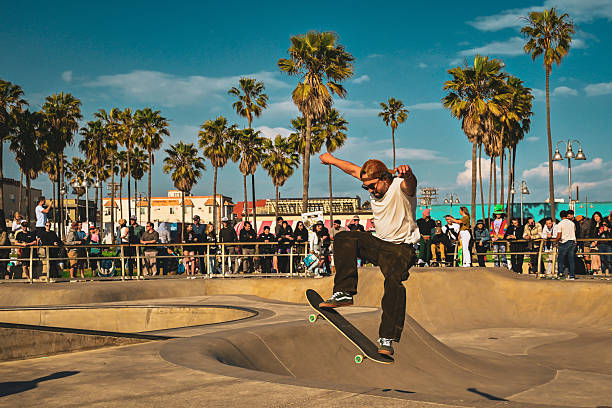A skateboarder performs a jump above the bowl at Venice Beach Skatepark surrounded by palm trees and a crowd of spectators in Los Angeles, California, United States