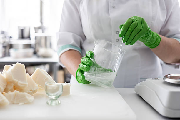 A laboratory technician in a white coat and green nitrile gloves stirs a liquid in a beaker. raft production of organic cosmetics involves weighing ingredients like raw shea butter on a digital scale