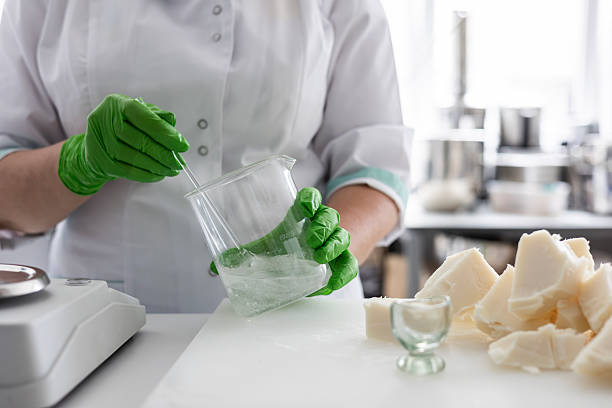 A laboratory technician in a white coat and green nitrile gloves stirs a liquid in a beaker. raft production of organic cosmetics involves weighing ingredients like raw shea butter on a digital scale