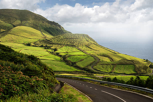 Winding asphalt road leads through lush green hills towards the coastline, under a partly cloudy sky in a peaceful rural landscape.