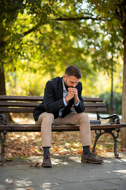 Thoughtful man sitting on a park bench, clasping his hands together and looking down, reflecting on personal or professional challenges.