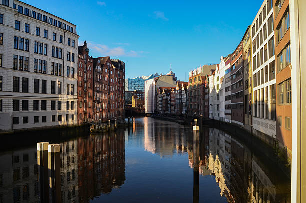 A picturesque view across one of Hamburg's historic canals (Fleet) in the city center. Traditional brick and modern buildings line the waterfront, reflecting beautifully in the dark water under a bright blue sky. In the background, the distinctive, wave-like roof of the modern Elbphilharmonie concert hall rises above the surrounding structures. This scene captures the unique blend of historic warehouse district architecture and modern cultural landmarks that defines Germany's second-largest city. The area is close to the UNESCO World Heritage site of Speicherstadt and the HafenCity district.