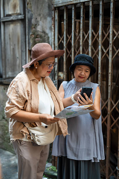 Senior asian women travelers checking map and phone together smiling and talking showing teamwork friendship and curiosity representing joyful retirement lifestyle exploration