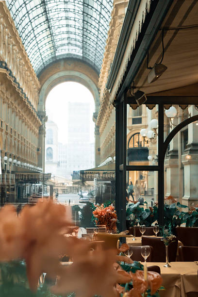 Atmospheric view inside the historic Galleria Vittorio Emanuele II in Milan, Italy. The image captures the elegant architecture, glass dome, and soft morning light filtering through the gallery, with a few tables and plants subtly visible in the foreground.