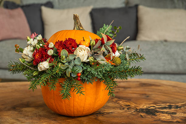 A vibrant floral arrangement inside a carved pumpkin, featuring red chrysanthemums, white roses, berries, and evergreen branches. The decorative centerpiece is displayed on a wooden table in a cozy indoor environment, perfect for autumn celebrations.