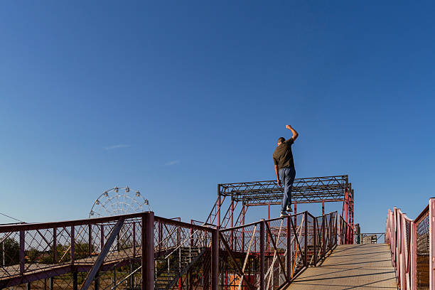 Freerunner walks across high railing in outdoor parkour structure, raising arm for balance against clear blue sky with Ferris wheel in distance. Precision in urban acrobatic movement. Parkour performance