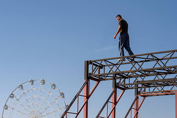 A freerunner walks on a narrow steel beam above the park, with a Ferris wheel in the background. The scene blends urban athleticism with a leisure park environment.