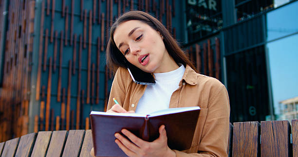 istock Close up of self employed female making phone call on mobile phone and taking notes while working remotely while sitting on bench about modern office building, outdoors 2245181730