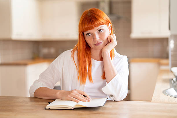 istock Young woman with red hair contemplates while writing in a notebook in a cozy kitchen setting 2245182512
