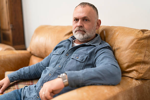 istock Middle-aged man sitting on a leather couch in a cozy room with a neutral background, looking thoughtfully at the camera after a long day 2245184487