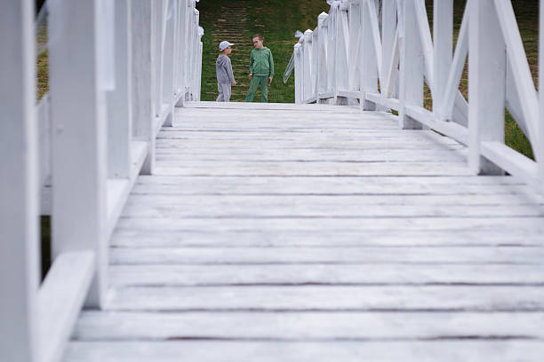 White wooden bridge creating a natural framing around two children standing on a grassy field ahead. Concept of perspective, outdoor childhood and calm summer mood.
