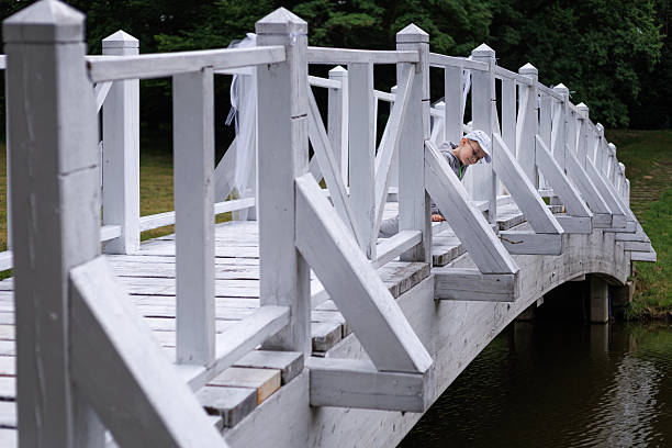 Child sitting on a white wooden bridge near the railing and looking outward over calm water. Concept of childhood curiosity, outdoor lifestyle and peaceful park atmosphere.