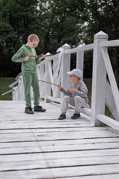Two young boys with fishing gear on a lakeside dock, examining their catch in a bucket. Perfect image depicting childhood curiosity and outdoor adventure.