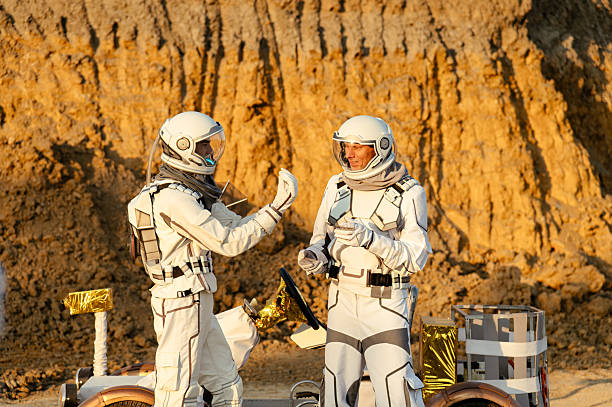 Two astronauts are standing on a lunar-like surface, engaged in a discussion while the setting sun casts a warm glow on the rocky terrain. They are near a rover designed for exploration.