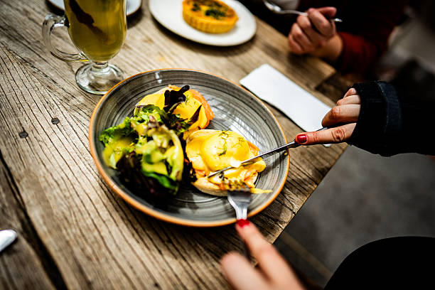 A mouthwatering close-up of Eggs Benedict served on a textured ceramic plate, featuring perfectly poached eggs with hollandaise sauce, accompanied by a side of fresh greens and drink. The image captures the moment just before eating, with a person's hands holding a fork, creating an inviting and appetizing scene.