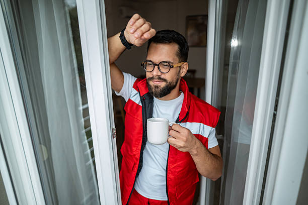 Male healthcare worker wearing a red vest and glasses, holding a coffee mug and looking thoughtfully out a window, taking a relaxing break from a demanding job