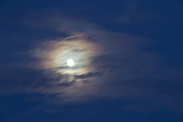 ethereal full moon shines through thin, fast-moving clouds, creating a stunning, iridescent corona effect against the dark blue night sky