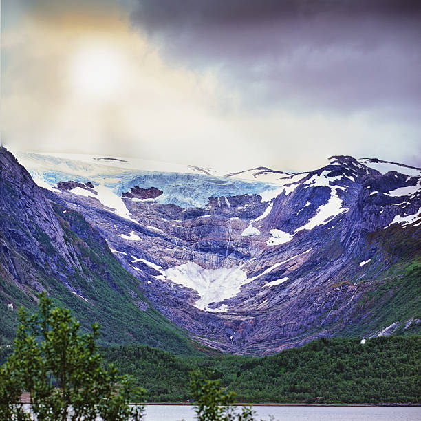 A breathtaking view of snow-covered glacier nestled in Norway's rugged mountains, surrounded by a lush green valley and a calm river, under an overcast sky with soft light.