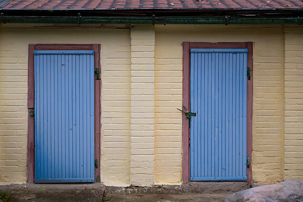 A pair of vertical blue wooden doors with red trim are set into a yellow-painted brick wall of an outbuilding. The symmetrical composition and weathered textures suggest rustic charm and utilitarian function. The concrete base and tiled roof add to the aged yet sturdy appearance.