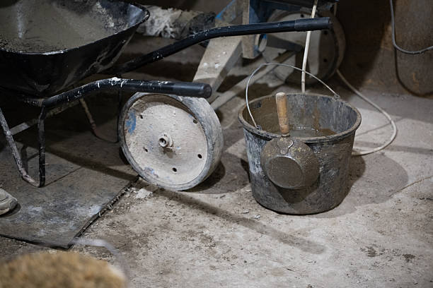 A worn metal bucket filled with wet mortar stands next to a wheelbarrow and trowel in a dusty indoor construction site. The rough concrete floor and scattered tools reflect a work-in-progress environment. The scene conveys manual labor, craftsmanship, and the gritty reality of building work.