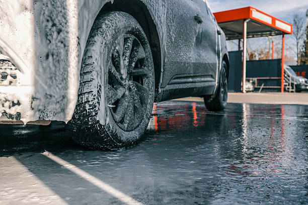 A close-up shot of a car wheel covered in soap suds at a car wash, with the wet ground reflecting the scene.
