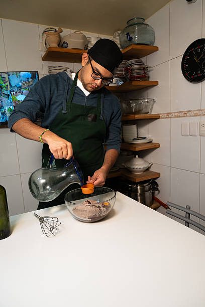 A Mexican chef pours water into a measuring cup, adding it to brownie mix and egg in a glass bowl during dessert prep in a traditional kitchen.