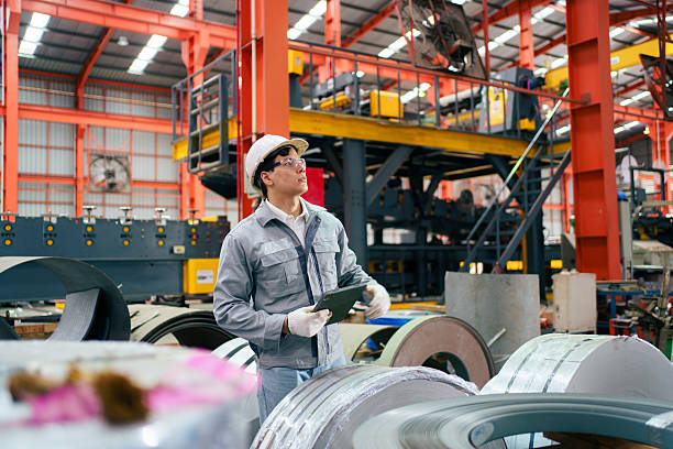 Latin male engineer wearing safety uniform and helmet holding digital tablet while inspecting machines and production equipment inside factory, representing engineering, and manufacturing process.