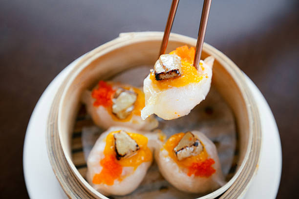 Close-up shot of chopsticks holding a gourmet dim sum topped with foie gras and orange fish roe, served in a bamboo steamer basket. The image highlights the elegance, texture, and luxury of Asian fine dining cuisine, captured under warm natural light.
