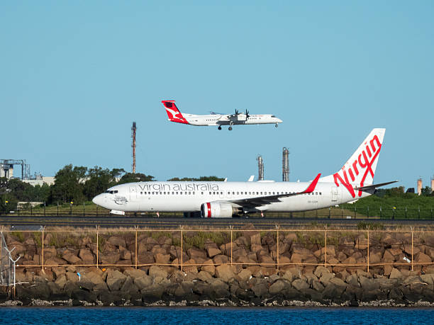 A Virgin Australia Boeing B737-8FE plane, registration VH-VUH, taxiing to the domestic terminal of Sydney Kingsford Smith Airport after landing as flight VA960 from Brisbane. In the background is a Qantas De Havilland Canada Dash 8-400 plane, registration VH-84G, landing on the third runway as flight QF2031 from Armidale. In the far background are smokestacks and factories in Botany. This image was taken from near Kyeemagh Beach, Botany Bay on a sunny afternoon on 6 November 2025.