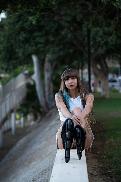 Young woman with tattoos and blue hair dye sitting on a wall, showing her inline skates after skating in the park