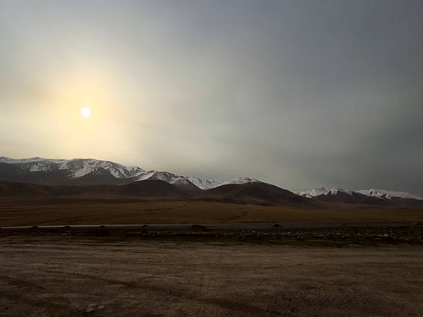 Low sun hanging above a wide valley framed by mountain ranges and open land in a remote region of Kyrgyzstan.