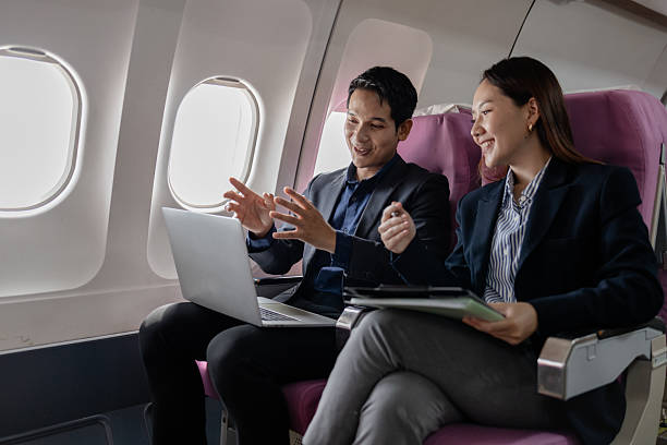istock Two Asian business professionals smiling and discussing ideas during a flight, sitting next to each other in an airplane cabin. The man gestures confidently while the woman listens attentively. 2245244616