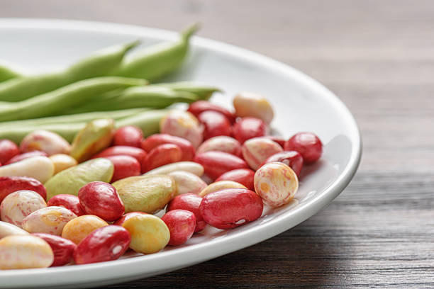 Closeup view of fresh green beans and colorful beans of different varieties and sizes on white plate on wooden table. Macro shot. Product of organic farming. Healthy eco vegetarian food.