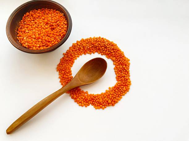 Top view of raw red lentils in a rustic clay bowl with a wooden spoon on a light background.