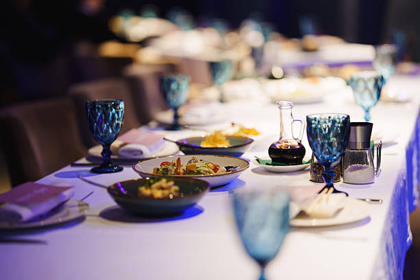 A long dining table is set with various dishes and turquoise glassware under soft lighting, creating an inviting atmosphere for a special evening meal.