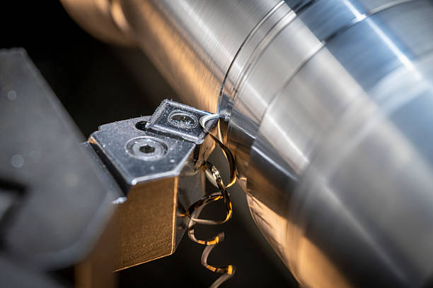 Close-up view of a lathe machine performing precision metalworking. The machine tool is cutting a cylindrical metal component, producing fine metal shavings as it operates.