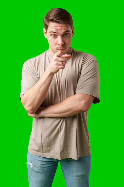 A young man with a serious expression pointing towards the viewer, standing against a bright green background