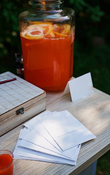 A bright drink station displays a large jar of orange beverage with sliced fruit. Nearby, blank postcards await writing, set on a wooden table in a lush green area.