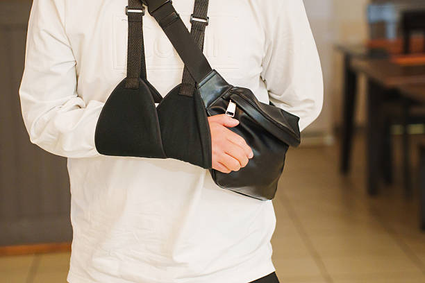 A person wearing a white long-sleeve shirt has their arm in a black sling while holding a small black bag. They are indoors, possibly in a cafe or home setting.