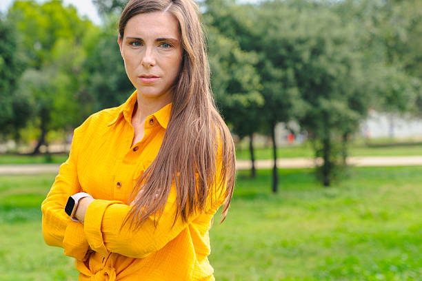 Confident woman wearing yellow shirt standing outdoors in green park looking at camera with copy space background, concept of leadership, determination, strength and professional lifestyle design use
