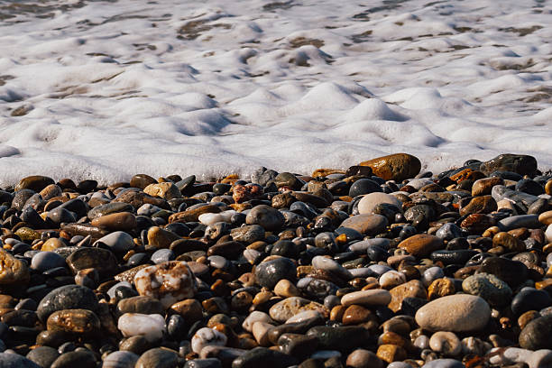 White sea foam washes over smooth, dark pebbles on a wild, natural beach shore. This close-up highlights the contrast in textures between the round stones and the frothy water