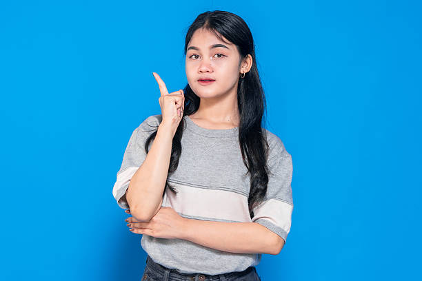 istock Young Asian woman with a thoughtful smile, pointing upward while standing against a blue background. She has long black hair and wears a casual gray t-shirt and black jeans. 2245289948