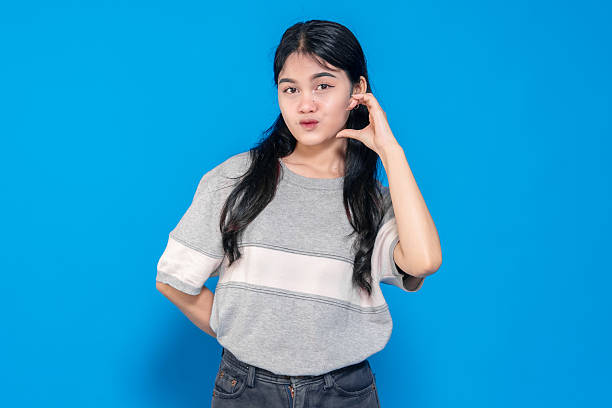 istock Young Asian woman making a playful pout expression while forming a heart shape with her fingers. She has long black hair, wears a casual gray t-shirt, and stands against a blue background. 2245289986