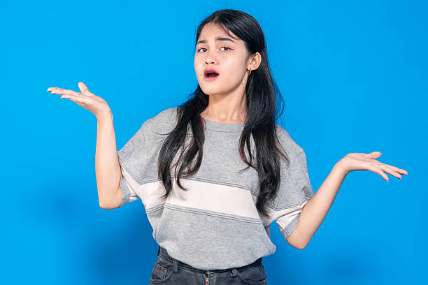 istock Young Asian woman with a confused expression, raising both hands in a questioning gesture. She looks uncertain or surprised, standing against a bright blue background. 2245290113
