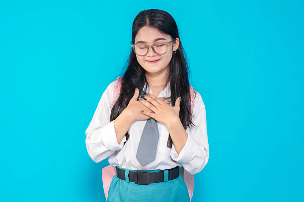 istock An Indonesian high school student in a school uniform with glasses and a backpack, smiling gently with closed eyes and hands on her chest, expressing gratitude against a bright blue background. 2245290825