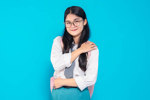 istock An Indonesian high school student in a school uniform, smiling gently while hugging herself in a comforting pose. She wears glasses and a backpack, standing against a bright blue background. 2245290838