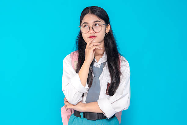 istock An Indonesian high school student in a school uniform with glasses and a backpack, displaying a thoughtful expression while resting her chin on her hand against a bright blue background. 2245290912