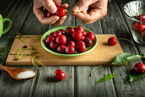 istock Hands remove stems from ripe cherries while a green bowl filled with the fruit sits on a wooden cutting board. Sugar and cherry leaves are nearby, adding to the scene 2245292974